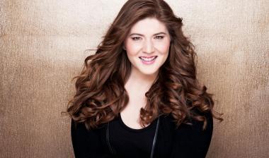 A headshot of Aria Umezawa. Smiling directly at the camera, Aria has long, curly, auburn hair and wears a black shirt in front a gradient bronze background.