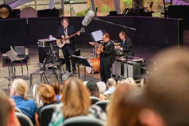 Members of the Brubeck Brothers Quartet performing bass, guitar, and drums in from of audience.