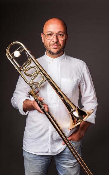 A bald man with light tan skin and a dark brown shadow of a beard, wearing glasses, a white button up shirt, and light jeans stares at the camera with a neutral expression. He holds a trombone with his right hand, his left in his pocket.