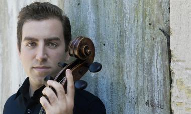 A white man with short brown hair wearing a collared navy blue shirt leans against a concrete wall holding the scroll of a cello.