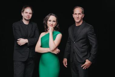 Two men in black suits stand on either side of a woman wearing a green dress with her hand to her chin.