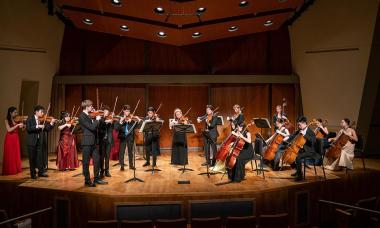 A group of string instrumentalists in black and red formalwear sit and stand on stage, playing