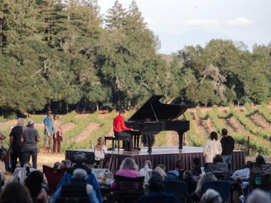 Hunter Noack plays piano in a field with audience members standing and seated in camping chairs while the sun sets