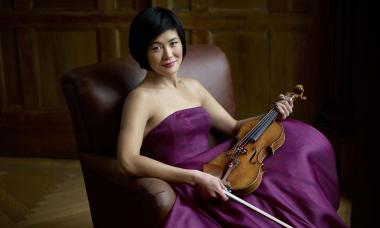 Jennifer Koh in a purple dress relaxing in a chair, holding her violin and bow