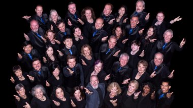 A group photo of The Choral Project’s 2024–25 ensemble, arranged in a dynamic, upward-facing formation against a black background. The singers, dressed in black with blue accents, smile warmly and extend one arm forward in a welcoming gesture. The group includes a diverse mix of ages, genders, and ethnicities, radiating unity and joy.