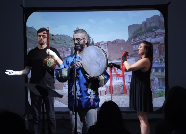 A singer holding a frame drum and two electronics musician standing in front of a projector screen.