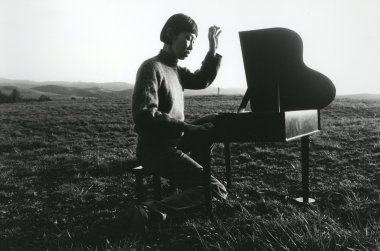 An Asian woman with a short bob sits at a small, toy piano in a field of grass with one hand raised as though she has just played a note