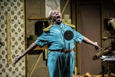 A man in a blue costume is singing in front of a microphone with his arms stretched out.