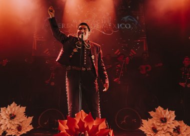 A man is dressed in traditional a Mexican mariachi attire standing in front of a mic with his hands up. There are flow appliqués at the bottom of the frame. 