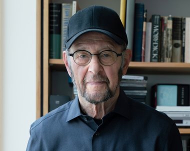 A man with a black baseball cap and glasses and black dress shirt is standing in front of a bookshelf. 