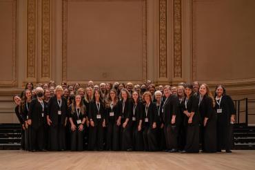 Group of fifty women smiling, wearing all black standing on the Carnegie Hall stage.