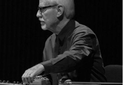John Bischoff is seated behind various instruments and electronics. He looks out of frame to the left in profile.