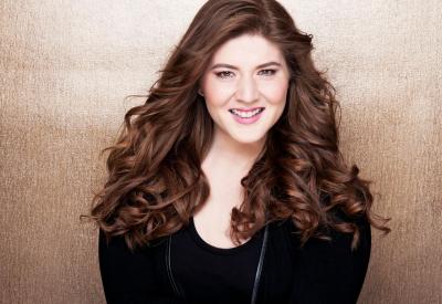 A headshot of Aria Umezawa. Smiling directly at the camera, Aria has long, curly, auburn hair and wears a black shirt in front a gradient bronze background.