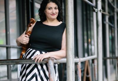 Wendy Richman holds her viola in her right arm as she leans against a wooden railing on a porch. She wears a black and white dress and smiles at the camera. 