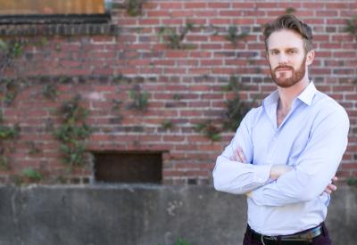 A white man with brownish ginger hair and beard wearing a button up shirt stands in front of a brick wall with vines climbing. His arms are crossed and his expression is soft.