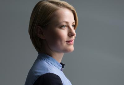 Portrait of a white woman with chin-length blonde hair, wearing a high necked light blue shirt. She is lightly smiling, nearly in side profile, with her face turned slightly  towards the camera.