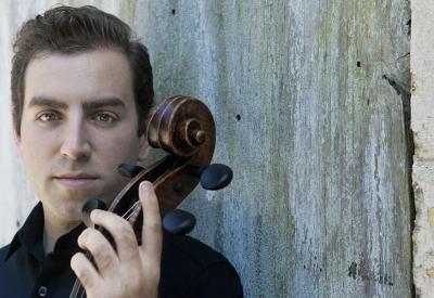 A white man with short brown hair wearing a collared navy blue shirt leans against a concrete wall holding the scroll of a cello.