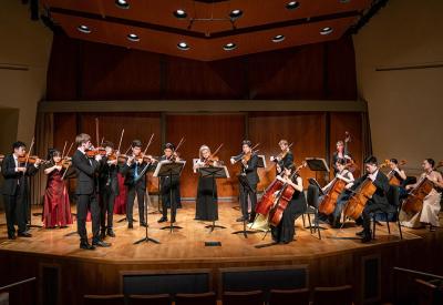A group of string instrumentalists in black and red formalwear sit and stand on stage, playing