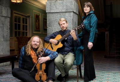 A woman, standing; a blonde man with a guitar, seated; a bearded man with red hair and a violin seated on the floor.