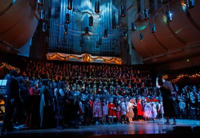 A choir of girls, young women, and non-binary singers hundreds strong perform onstage at a festively decorated Davies Symphony Hall, conducted by Valérie Sainte-Agathe, in low blue lighting.