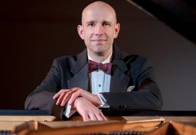 Christopher Taylor in a suit leaning on his piano looking at the camera. 