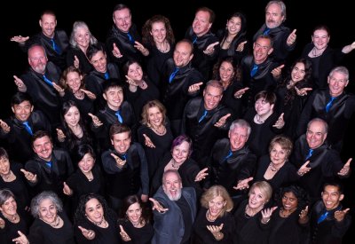 A group photo of The Choral Project’s 2024–25 ensemble, arranged in a dynamic, upward-facing formation against a black background. The singers, dressed in black with blue accents, smile warmly and extend one arm forward in a welcoming gesture. The group includes a diverse mix of ages, genders, and ethnicities, radiating unity and joy.