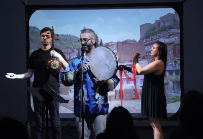 A singer holding a frame drum and two electronics musician standing in front of a projector screen.