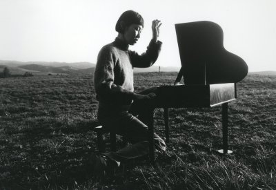 An Asian woman with a short bob sits at a small, toy piano in a field of grass with one hand raised as though she has just played a note