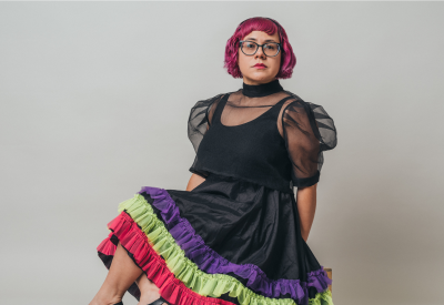 A woman with short, hot pink hair and glasses poses, seated in a black dress with a colorful trim.