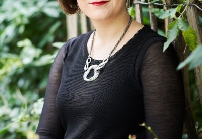 A women in black formal attire stands in front of greenery. 