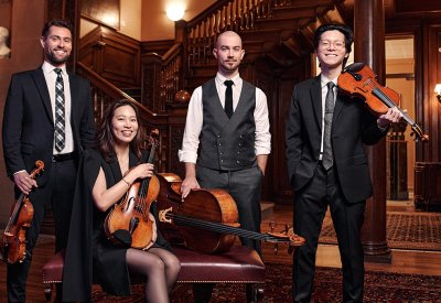 Portrait of the Dover Quartet standing in front of a wooden staircase. All of the members are by their respective instrument