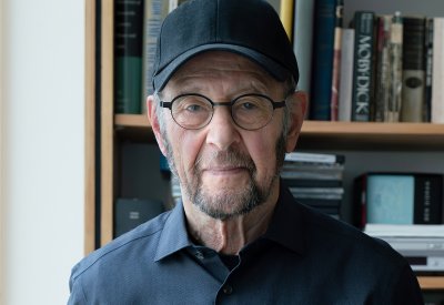 A man with a black baseball cap and glasses and black dress shirt is standing in front of a bookshelf. 